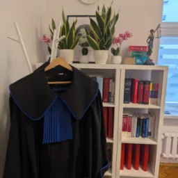 Toga adwokacka z niebieskim żabotem na wieszaku, na tle białego regału z książkami prawniczymi, figurką Temidy i zegarem ściennym.