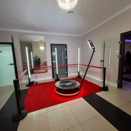George Studio Kreatywne - Rotacyjna fotobudka 360 z czerwonym dywanem i oświetleniem LED, widok z góry. W tle lustro, w którym odbijają się dwie osoby. Eleganckie wnętrze.