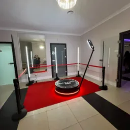 Rotacyjna fotobudka 360 z czerwonym dywanem i oświetleniem LED, widok z góry. W tle lustro, w którym odbijają się dwie osoby. Eleganckie wnętrze.