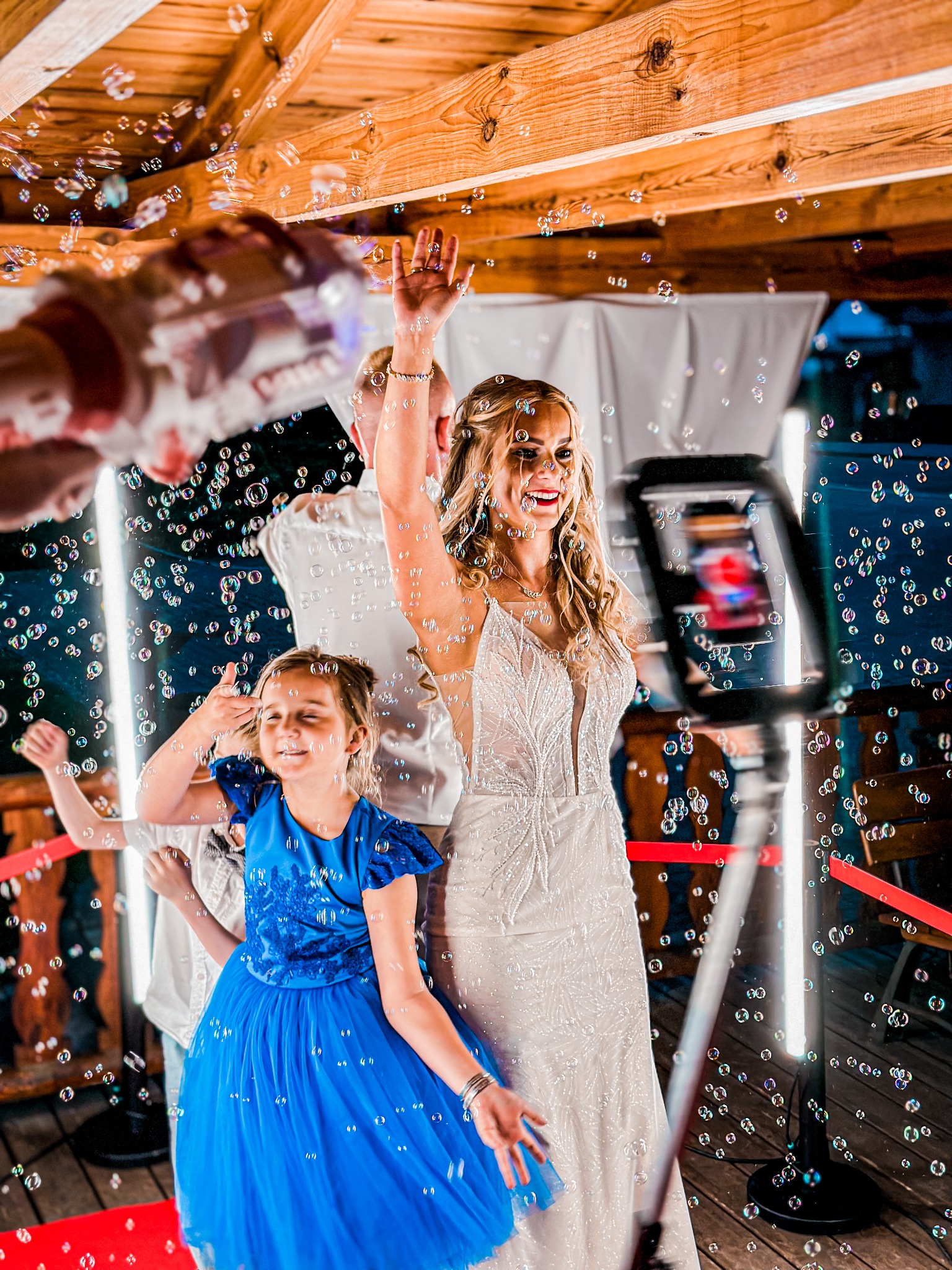 Uśmiechnięta panna młoda i dziewczynka w niebieskiej sukience pozują w chmurze baniek mydlanych przed fotobudką we Wrocławiu, widoczny czerwony dywan i drewniana konstrukcja.