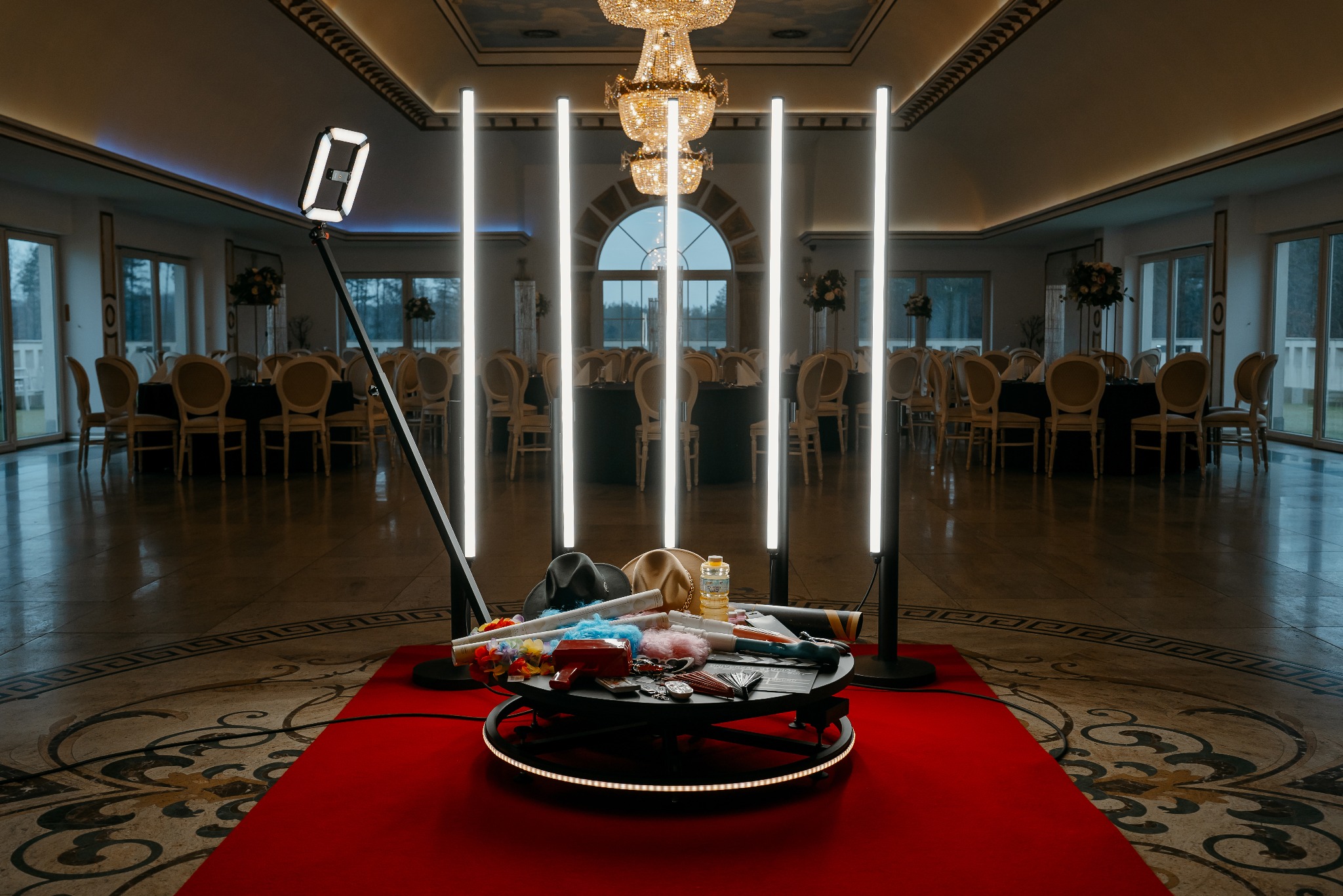 Fotobudka 360 stopni z rekwizytami na czerwonym dywanie w eleganckiej sali balowej z kryształowym żyrandolem i stołami dla gości.