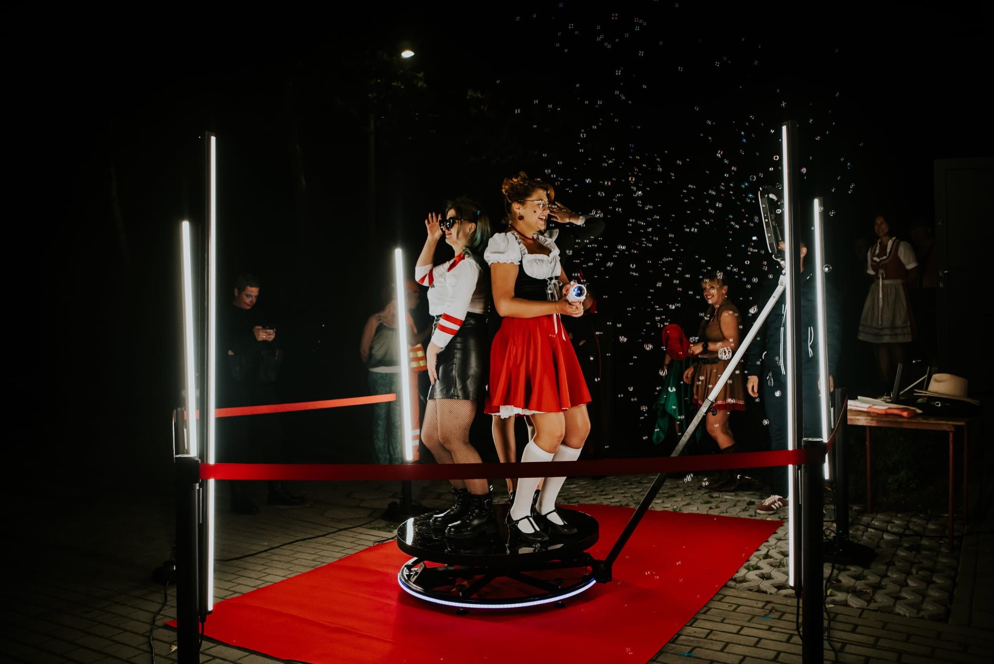 Dwie uśmiechnięte kobiety w strojach stylizowanych na bawarskie, pozują na obrotowej platformie otoczonej ledowymi lampami i czerwoną taśmą, w tle widoczne bańki mydlane i inne osoby.