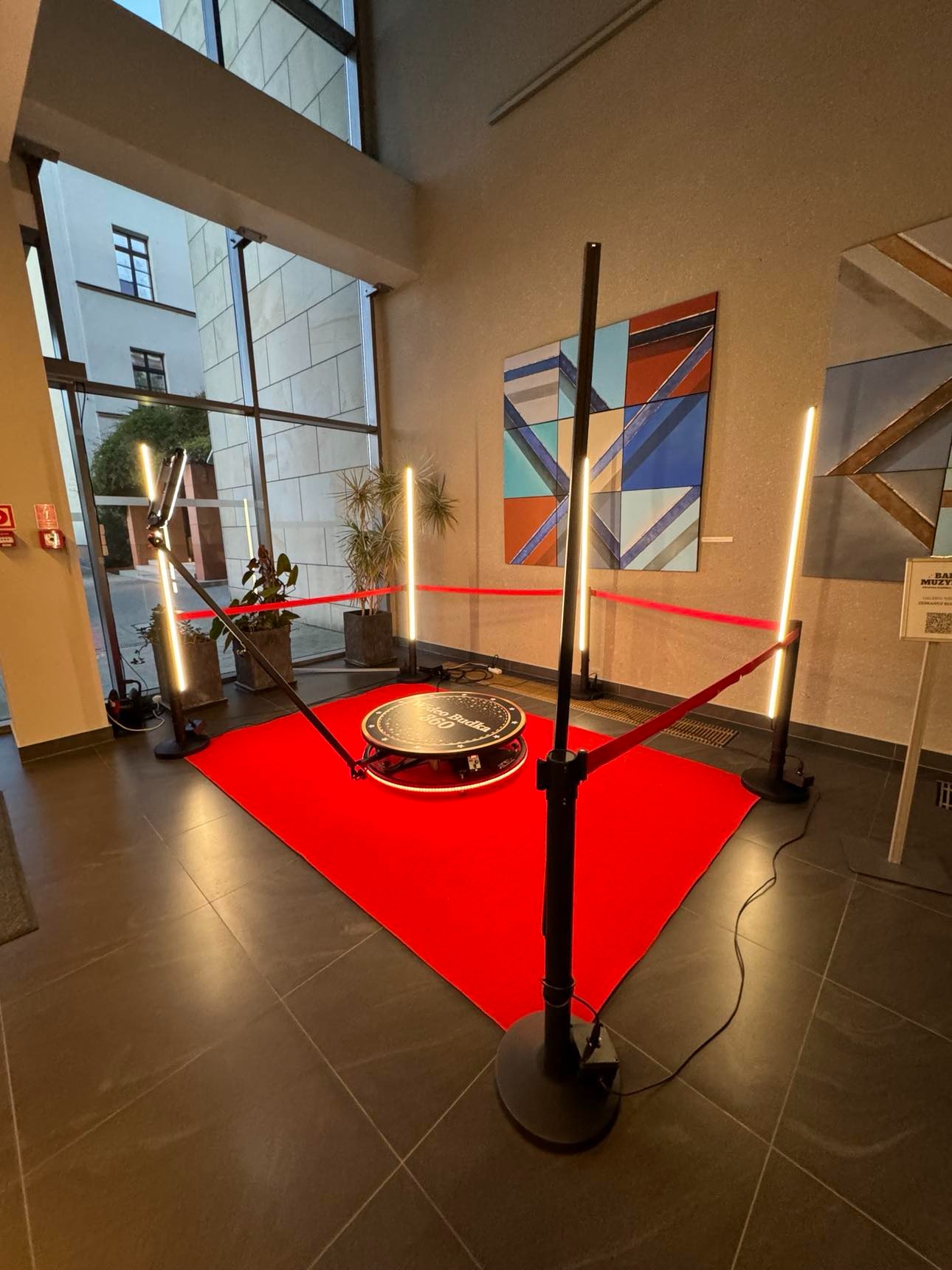 Oświetlona platforma obrotowa 360 stopni na czerwonym dywanie, otoczona barierkami z czerwonej taśmy, ustawiona w przestronnym holu z dużymi oknami i nowoczesnymi obrazami na ścianach.