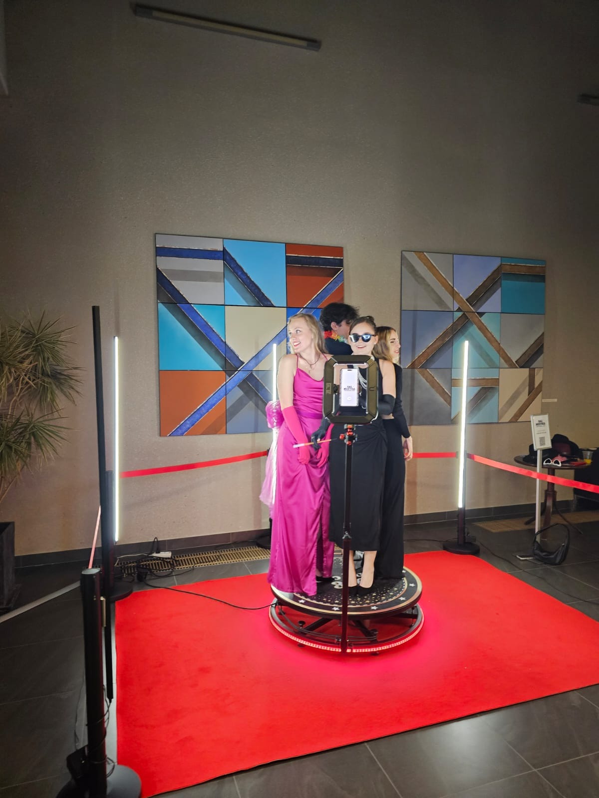 Trzy osoby w eleganckich strojach pozują na obrotowej platformie fotobudki 360 na czerwonym dywanie przed ścianą z abstrakcyjnymi obrazami.