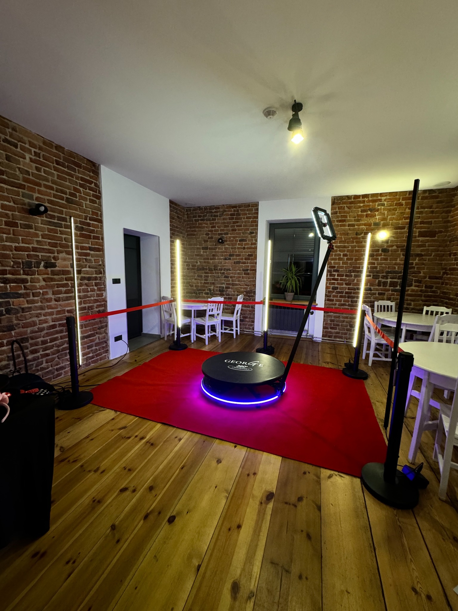 Rotacyjna fotobudka 360 z podświetleniem LED na czerwonym dywanie w pomieszczeniu z ceglanymi ścianami i drewnianą podłogą, oświetlona lampami LED.