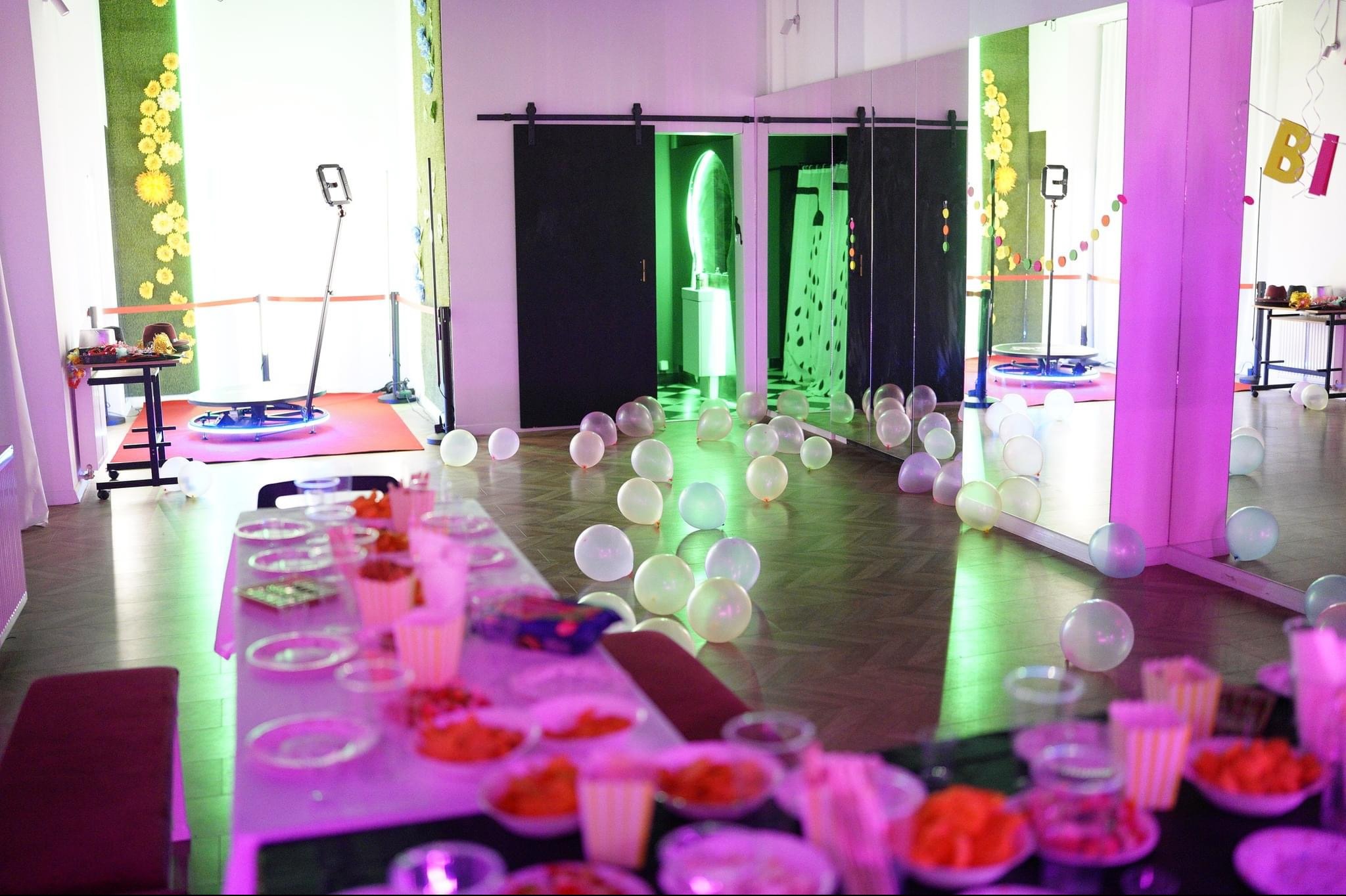 Sala udekorowana na imprezę z fotobudką 360, balonami na podłodze, stolikiem z przekąskami i lustrami.