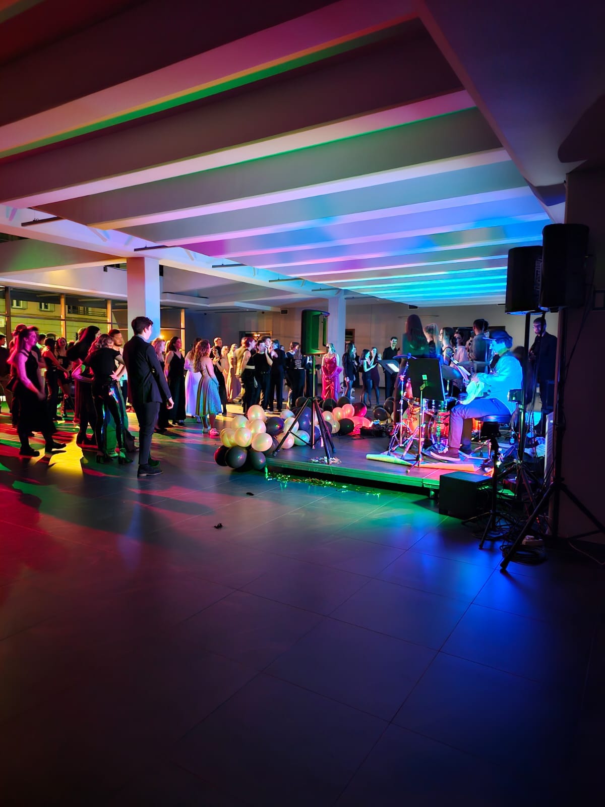Sala balowa z zespołem muzycznym na scenie, tancerze i goście w tle, dekoracja balonowa w odcieniach bieli i czerni, oświetlenie sceniczne w kolorach tęczy.