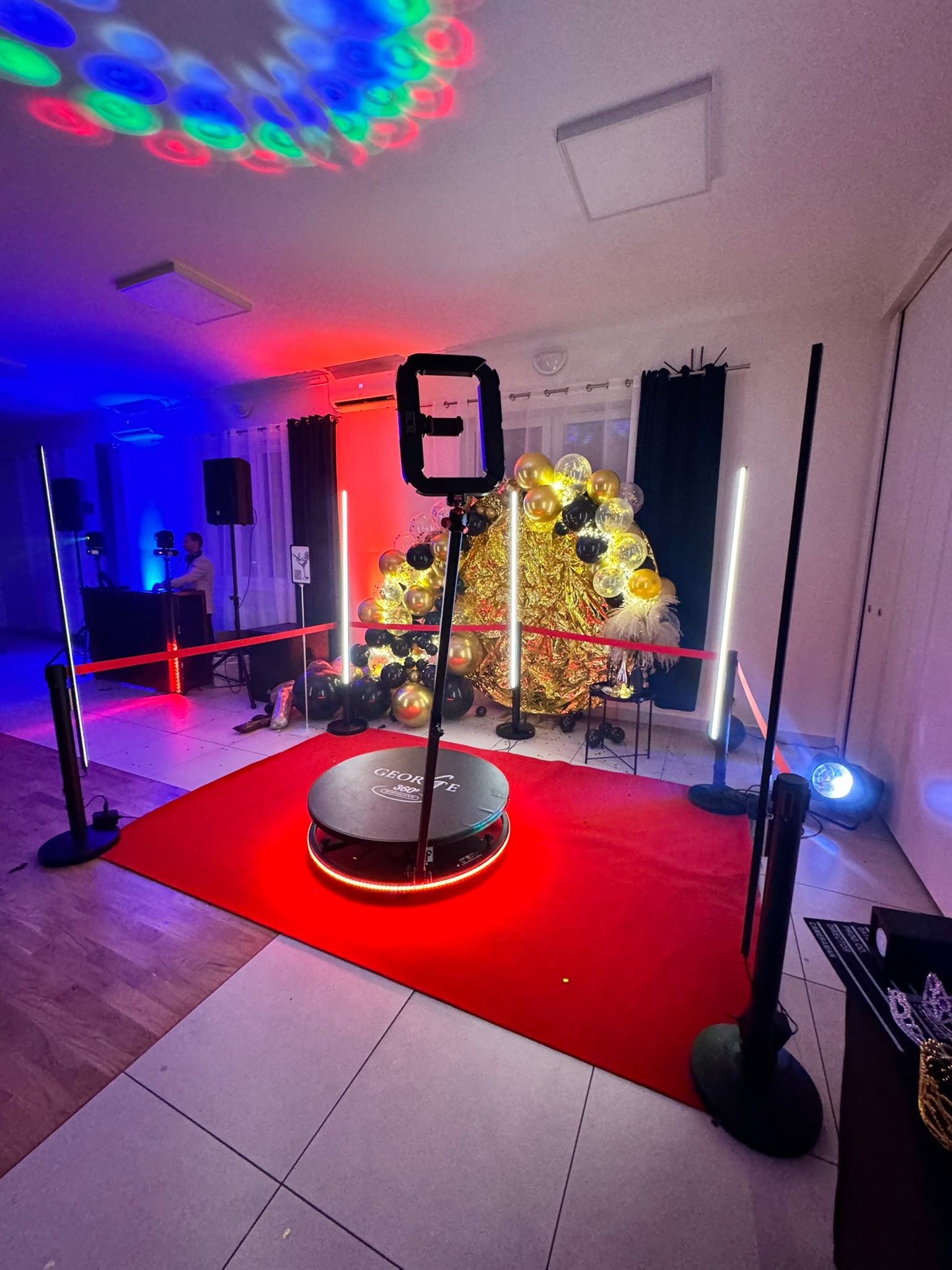Świetlna dekoracja imprezowa z obrotową platformą, czerwonym dywanem i balonami w kolorach złota i czerni. W tle DJ i efekty świetlne.