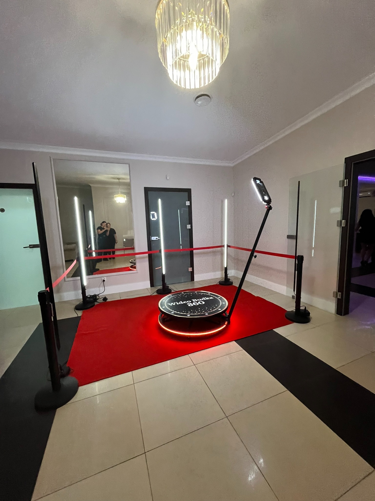 Rotacyjna fotobudka 360 z czerwonym dywanem i oświetleniem LED, widok z góry. W tle lustro, w którym odbijają się dwie osoby. Eleganckie wnętrze.