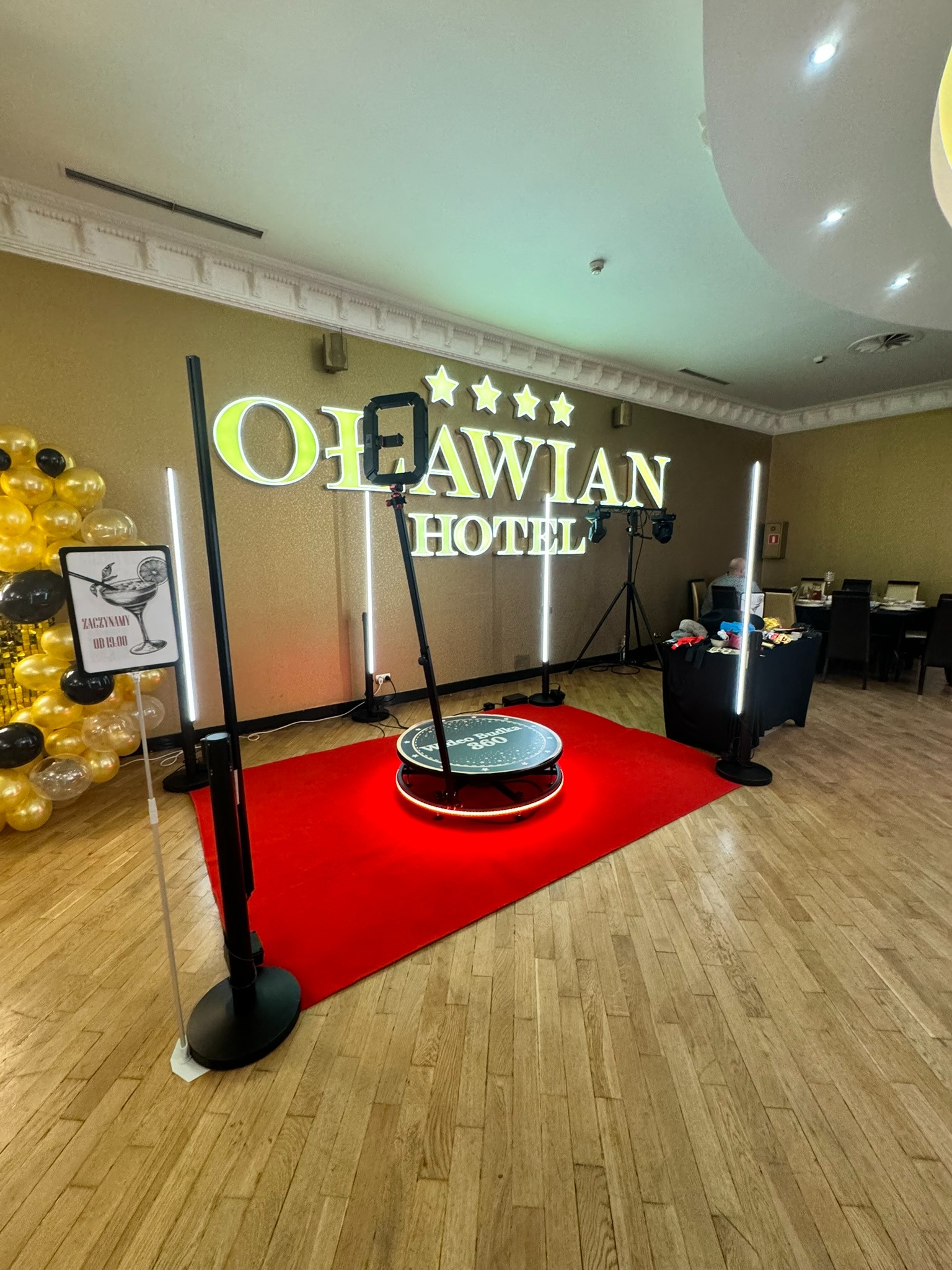 Fotobudka 360 w hotelu Otavian. Platforma z oświetleniem na czerwonym dywanie, dekoracje balonowe i szyld informujący o rozpoczęciu imprezy.