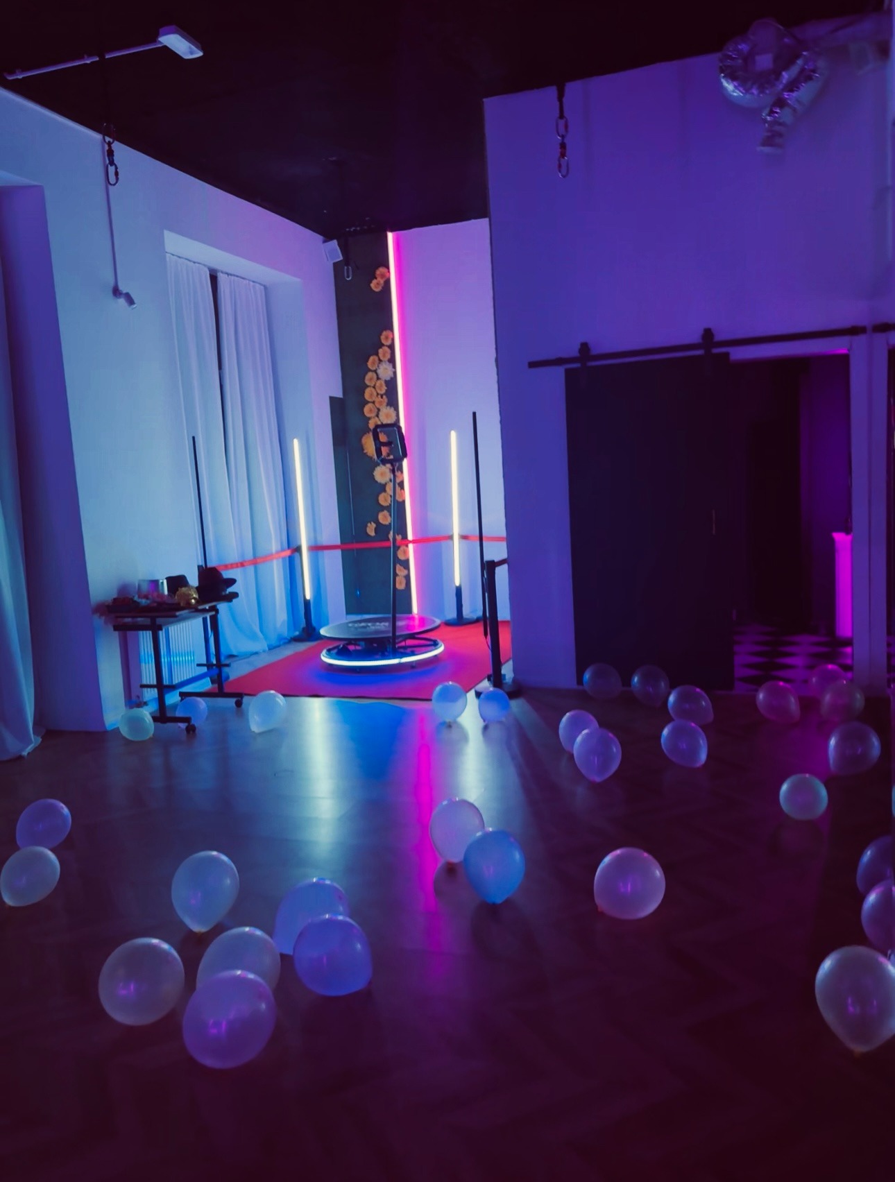 Oświetlona platforma 360° z neonami i czerwonym dywanem, otoczona rozrzuconymi balonami na drewnianej podłodze, w pomieszczeniu z dekoracjami kwiatowymi.