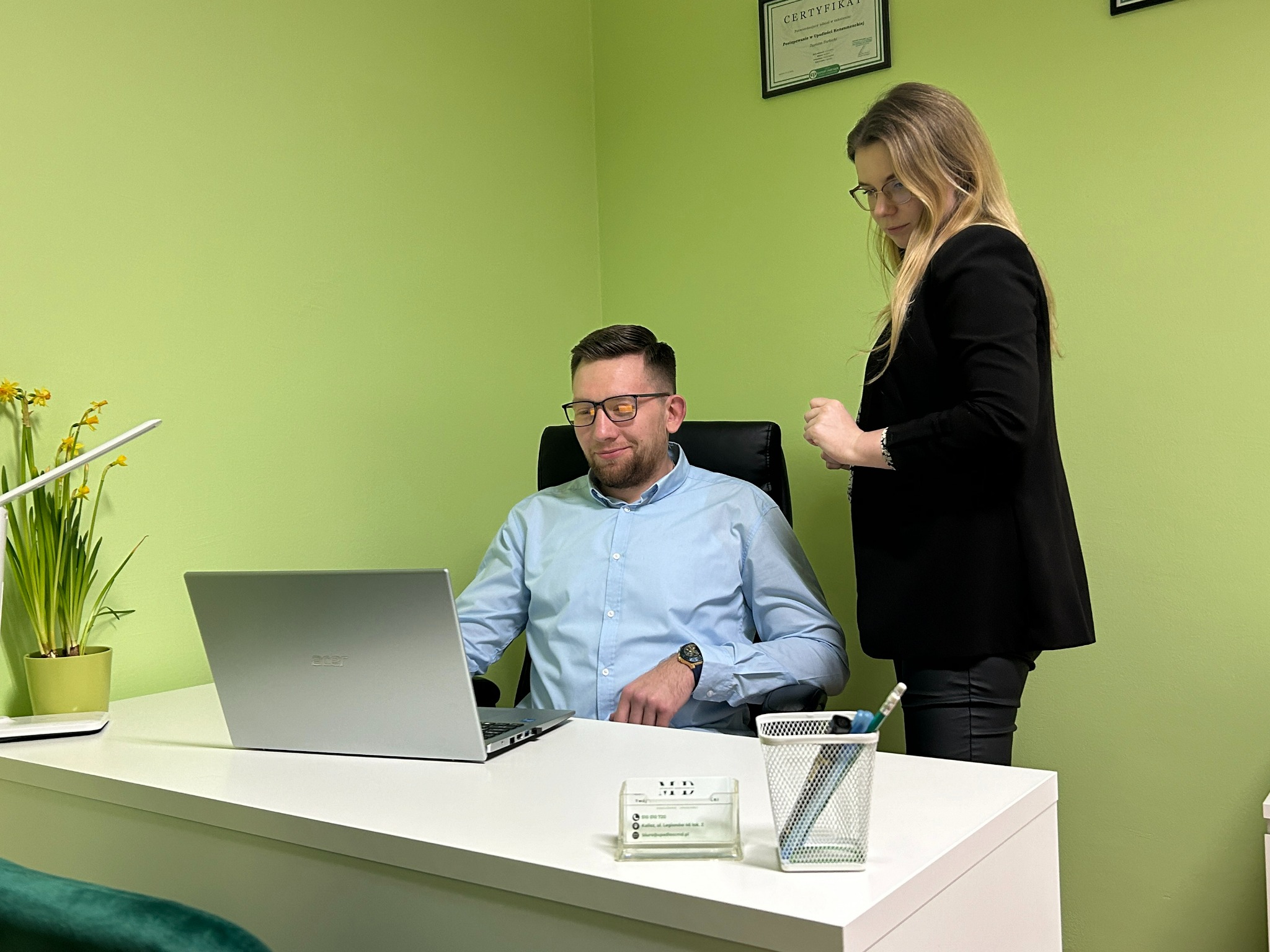 Konsultacja prawna: mężczyzna przy laptopie, kobieta obok, certyfikat na ścianie, biuro w odcieniach zieleni. Profesjonalne doradztwo w sprawach upadłości konsumenckiej.