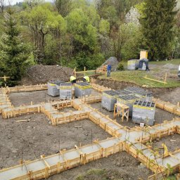 Idea Budownictwo - Wylane betonowe fundamenty w drewnianym szalunku na placu budowy, widoczne zbrojenia, zapasy bloczków styrobud, pracownicy w odblaskowych kamizelkach i ciężarówka w tle, teren pagórkowaty.