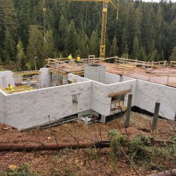 Idea Budownictwo - Budowa domu jednorodzinnego w stanie surowym otwartym, ściany z bloczków, drewniane szalunki stropu, widoczny żuraw budowlany i zalesione wzgórze w tle.