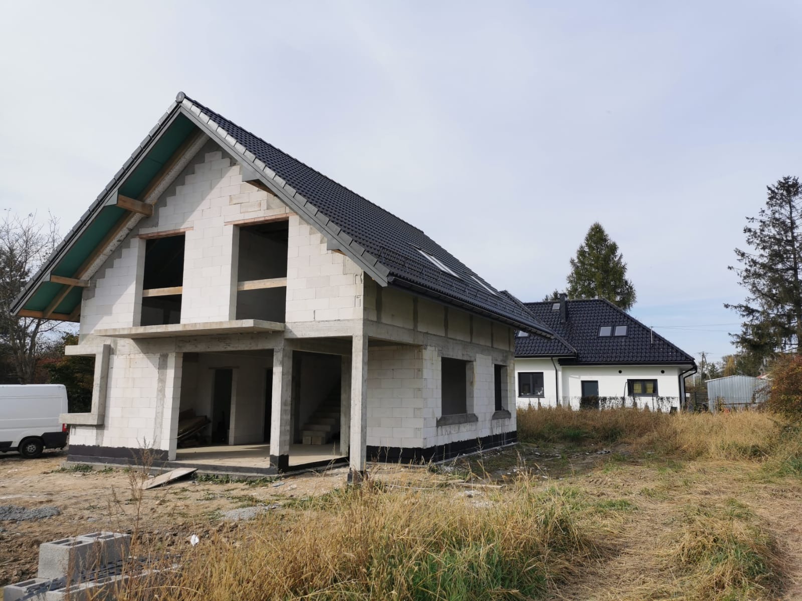 Surowy stan domu murowanego z białych bloczków, widoczne niezabudowane otwory okienne i drzwiowe, czarny dach, w tle drugi, wykończony dom.
