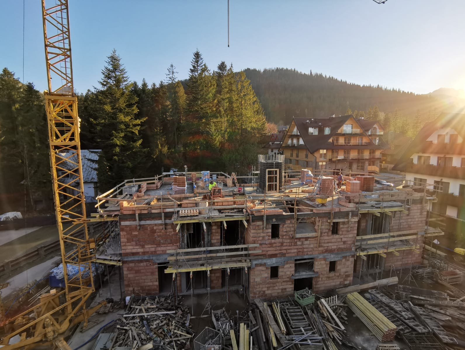 Wznoszenie dwukondygnacyjnego budynku z czerwonej cegły na tle górskiego krajobrazu, widoczny żółty dźwig i pracujący robotnicy.