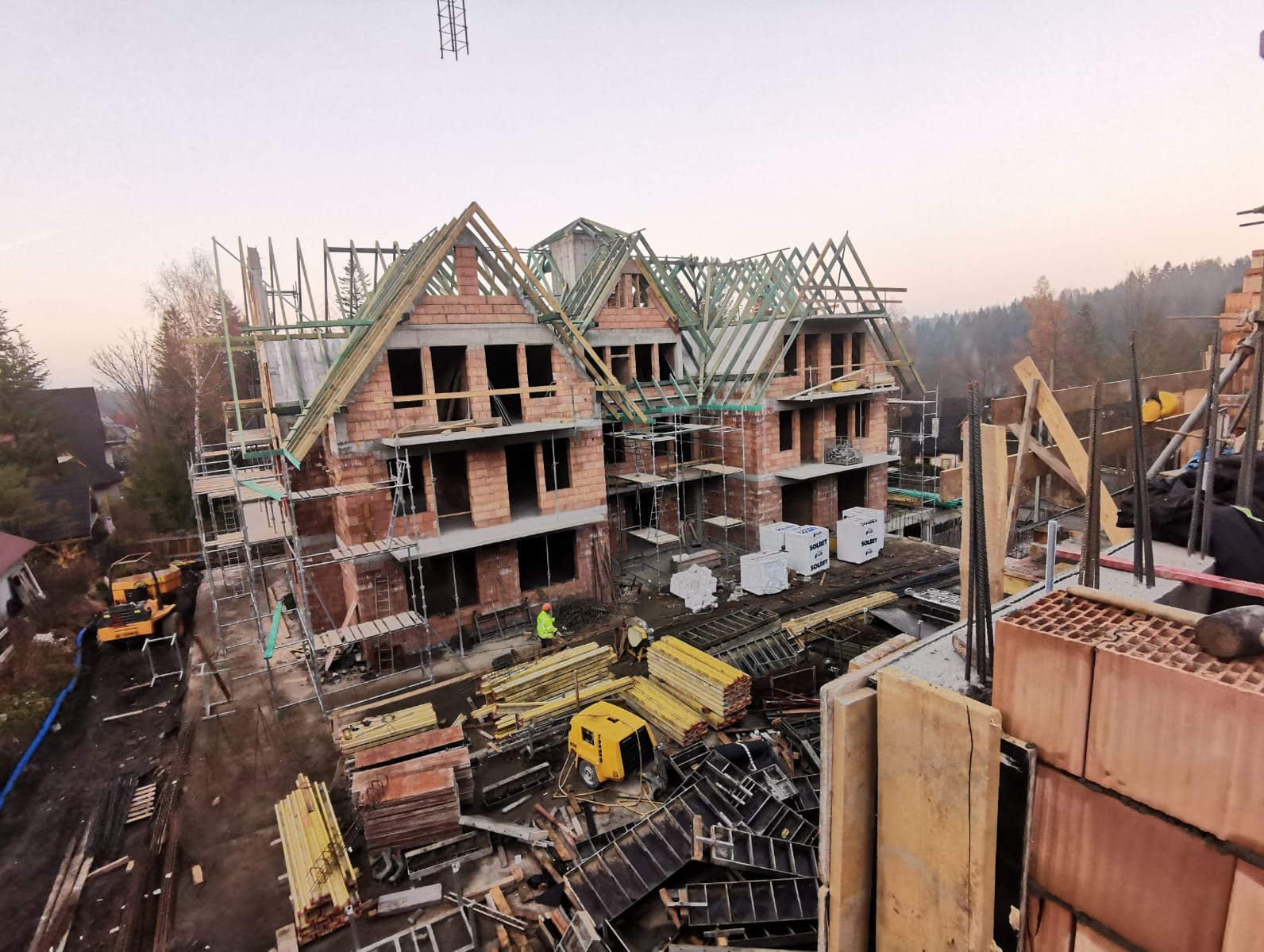 Wysoki kąt widzenia na budowę trzech domów murowanych w stanie surowym otwartym, z widocznymi rusztowaniami, materiałami budowlanymi i pracownikiem w odblaskowej kamizelce przy jednym z budynków.