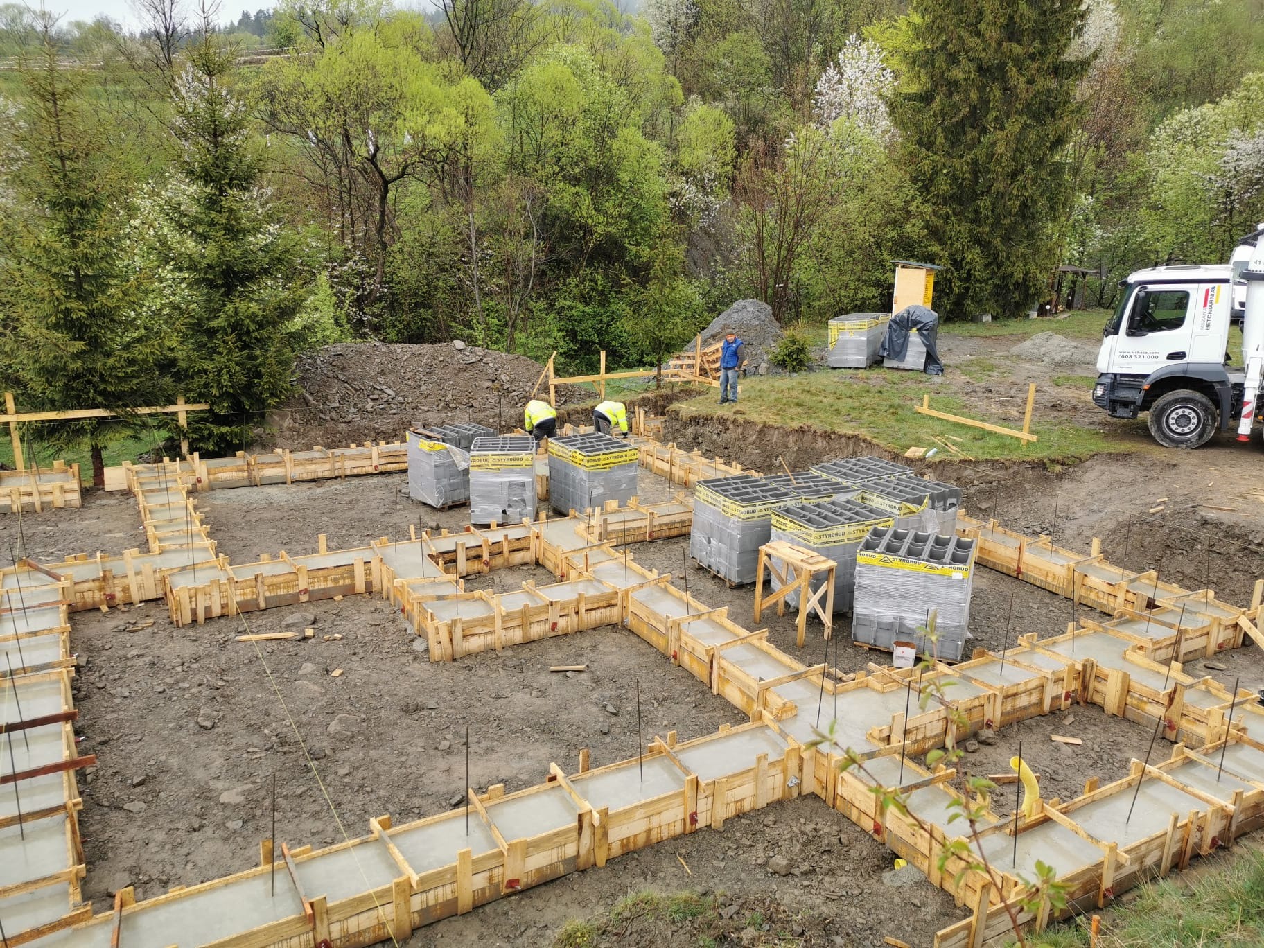 Wylane betonowe fundamenty w drewnianym szalunku na placu budowy, widoczne zbrojenia, zapasy bloczków styrobud, pracownicy w odblaskowych kamizelkach i ciężarówka w tle, teren pagórkowaty.