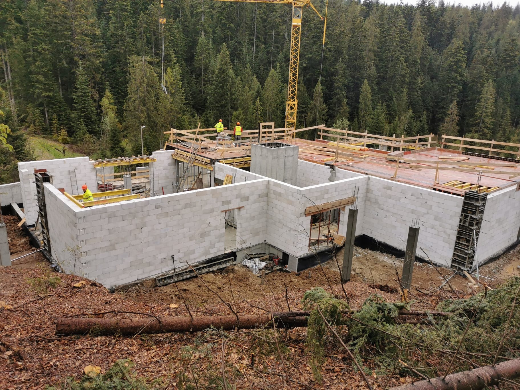 Budowa domu jednorodzinnego w stanie surowym otwartym, ściany z bloczków, drewniane szalunki stropu, widoczny żuraw budowlany i zalesione wzgórze w tle.