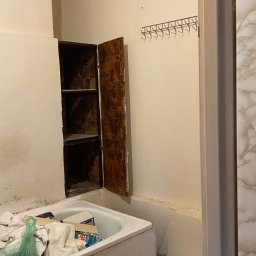 Perfectfix Remont - Zaniedbana łazienka z otwartą szafką w ścianie, białą wanną wypełnioną przedmiotami i rurami na ścianie. Widoczne ślady wilgoci i brak estetyki.