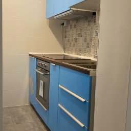 Perfectfix Remont - Błękitna zabudowa kuchenna z piekarnikiem i płytą grzewczą, wykończona brązowym blatem. Nad nią okap i dekoracyjne płytki. Szare płytki podłogowe.