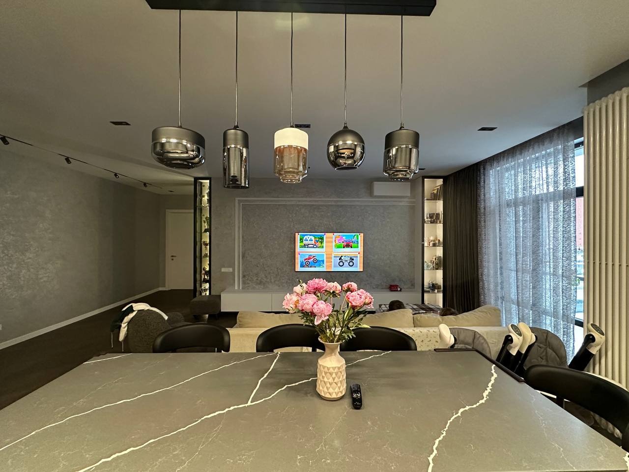 Nowoczesny salon z szarą ścianą, marmurowym stołem, bukietem różowych piwonii w ceramicznym wazonie i designerskimi lampami wiszącymi nad stołem.