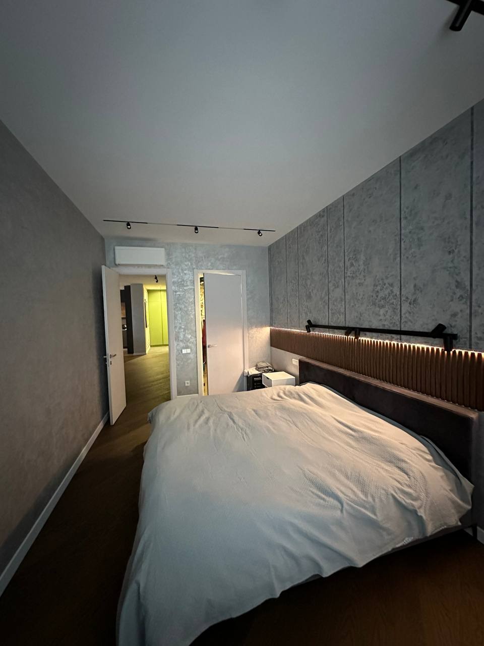 Sypialnia z łóżkiem, ściany wykończone panelami dekoracyjnymi o fakturze betonu, sufit z oświetleniem szynowym, widok na otwarte drzwi do kolejnego pomieszczenia.