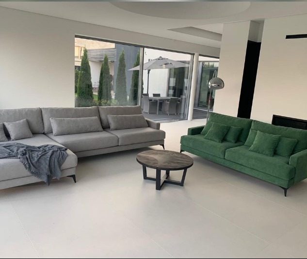 Jasne wnętrze salonu z szarym narożnikiem z pledem, zieloną sofą i okrągłym stolikiem kawowym na tle panoramicznego okna z widokiem na ogród z parasolem.