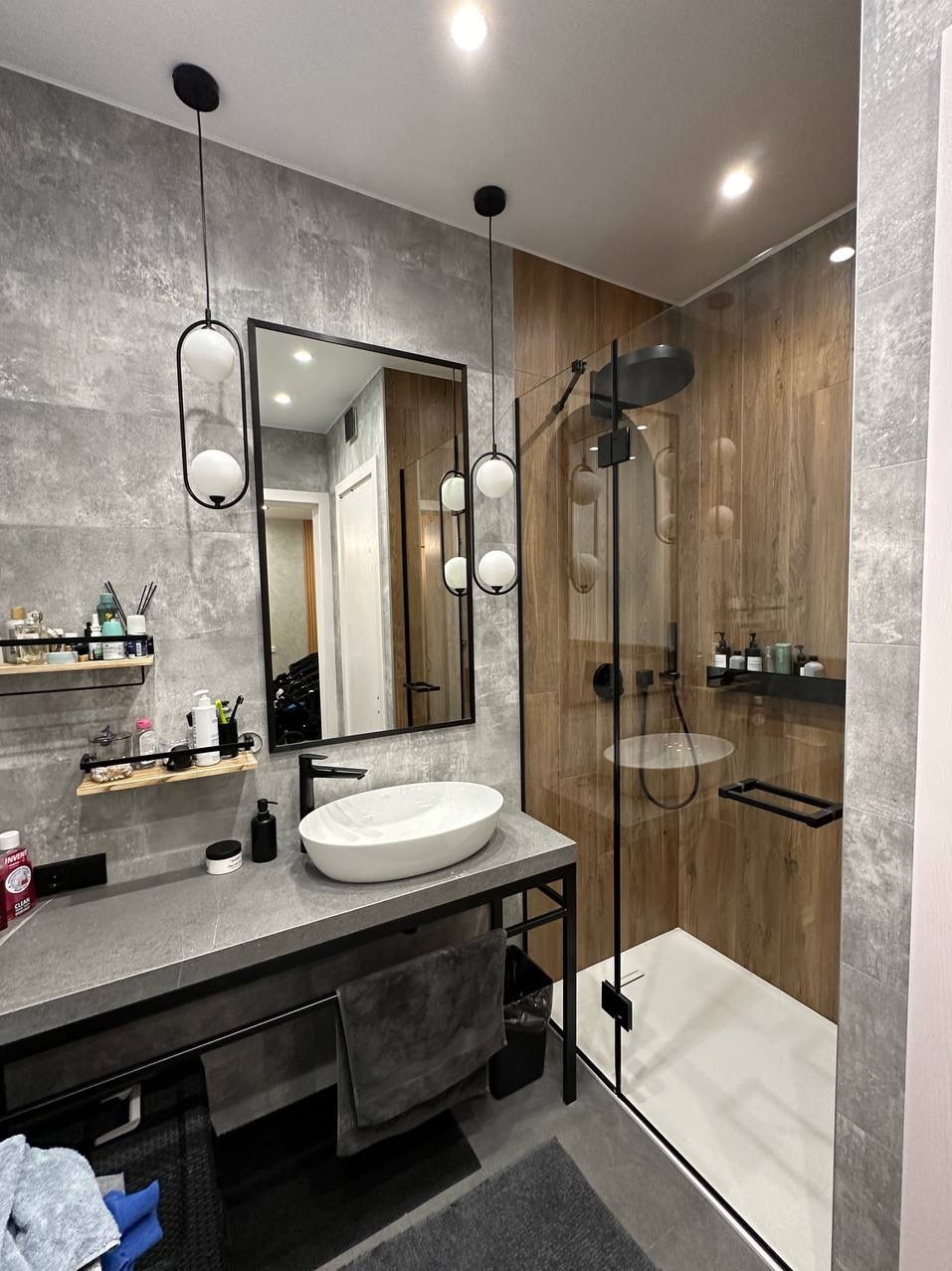 Nowoczesna łazienka z szarą, betonową umywalką na metalowym stelażu, owalną białą umywalką nablatową, czarną armaturą, lustrem w czarnej ramie, wiszącymi lampami z białymi kulami i kabiną...