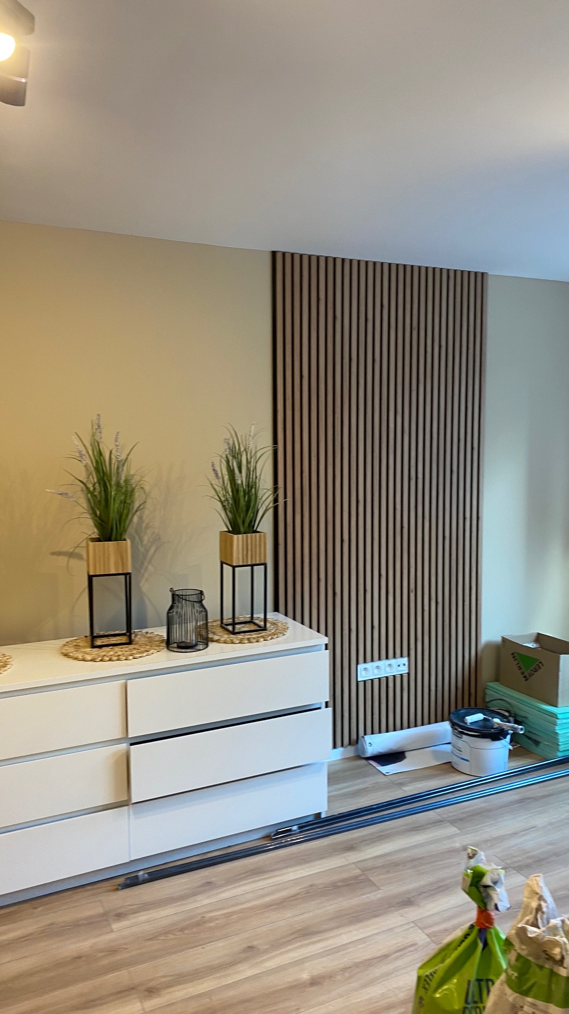 Biała komoda z dekoracjami roślinnymi i drewniana ściana z lameli w tle. Widoczne materiały budowlane sugerują prace wykończeniowe.