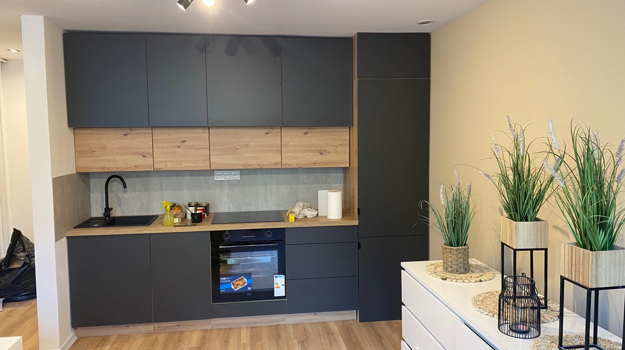 Nowoczesna kuchnia z szarymi szafkami i drewnianymi akcentami. Na blacie kuchennym widoczne akcesoria, a obok dekoracyjne rośliny w donicach. Jasna podłoga i ściany.