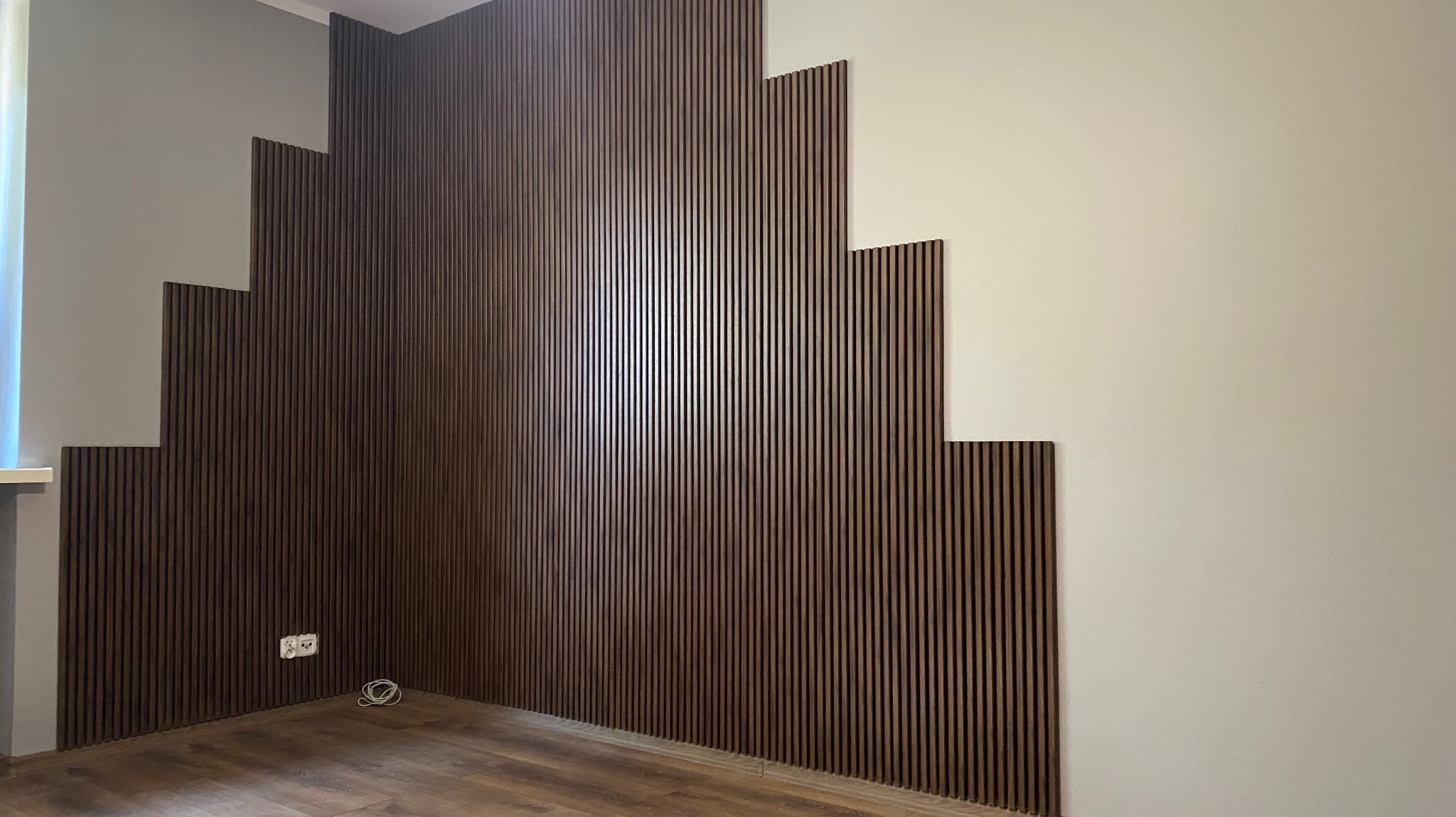 Brązowe lamele dekoracyjne na ścianie, ułożone schodkowo, tworzące nowoczesny design wnętrza. Widoczne gniazdko elektryczne i fragment podłogi.