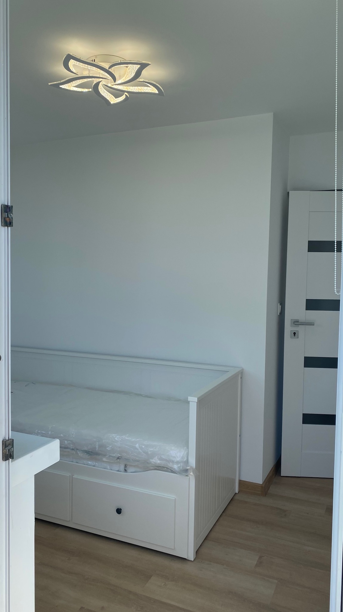 Jasny pokój z białym łóżkiem z szufladami, podłogą imitującą drewno i nowoczesną lampą sufitową w kształcie kwiatu. Widok zza otwartych drzwi.