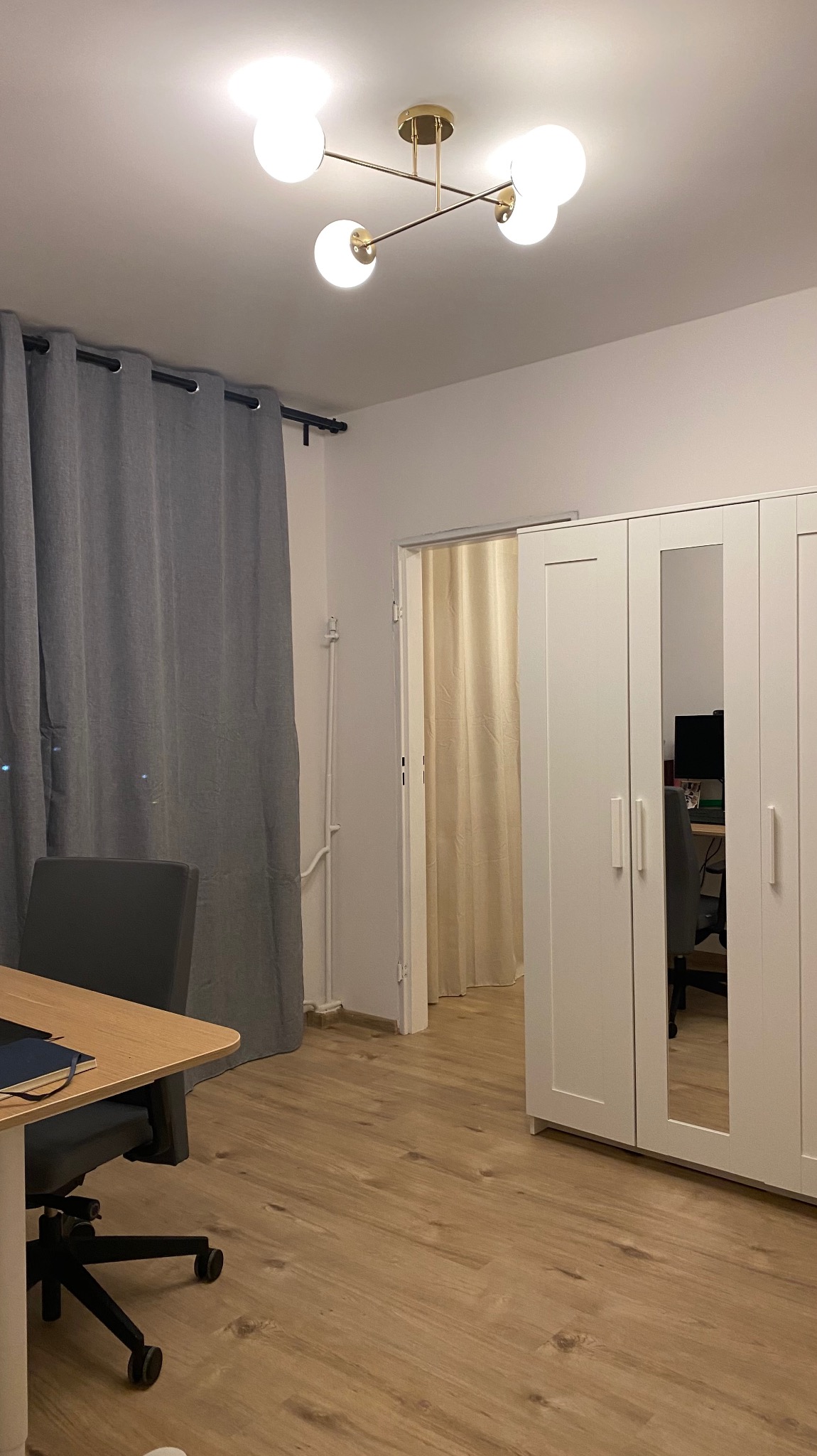 Jasne wnętrze pokoju z drewnianą podłogą, szarą zasłoną, białymi szafami i nowoczesną lampą sufitową z kulistymi kloszami. Widok na sąsiednie pomieszczenie.