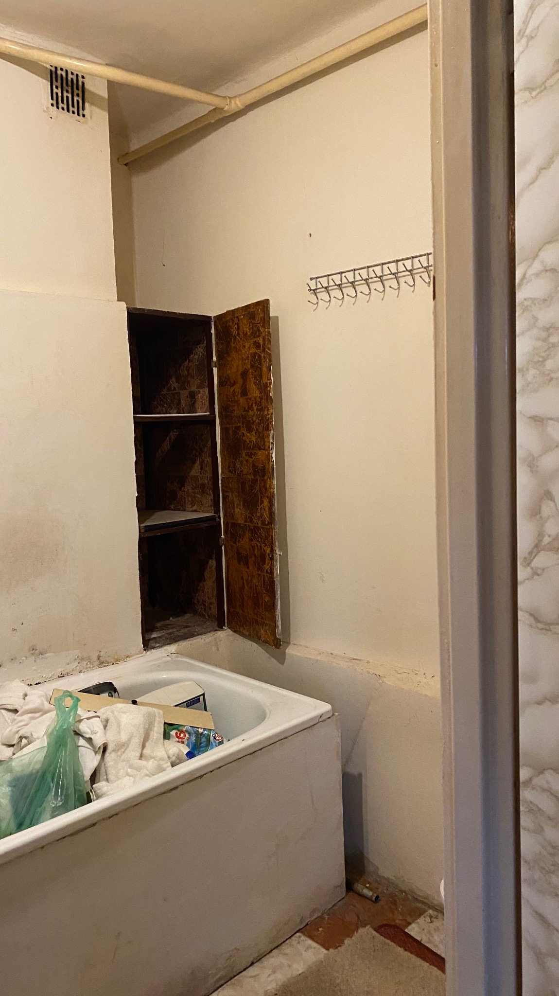 Zaniedbana łazienka z otwartą szafką w ścianie, białą wanną wypełnioną przedmiotami i rurami na ścianie. Widoczne ślady wilgoci i brak estetyki.