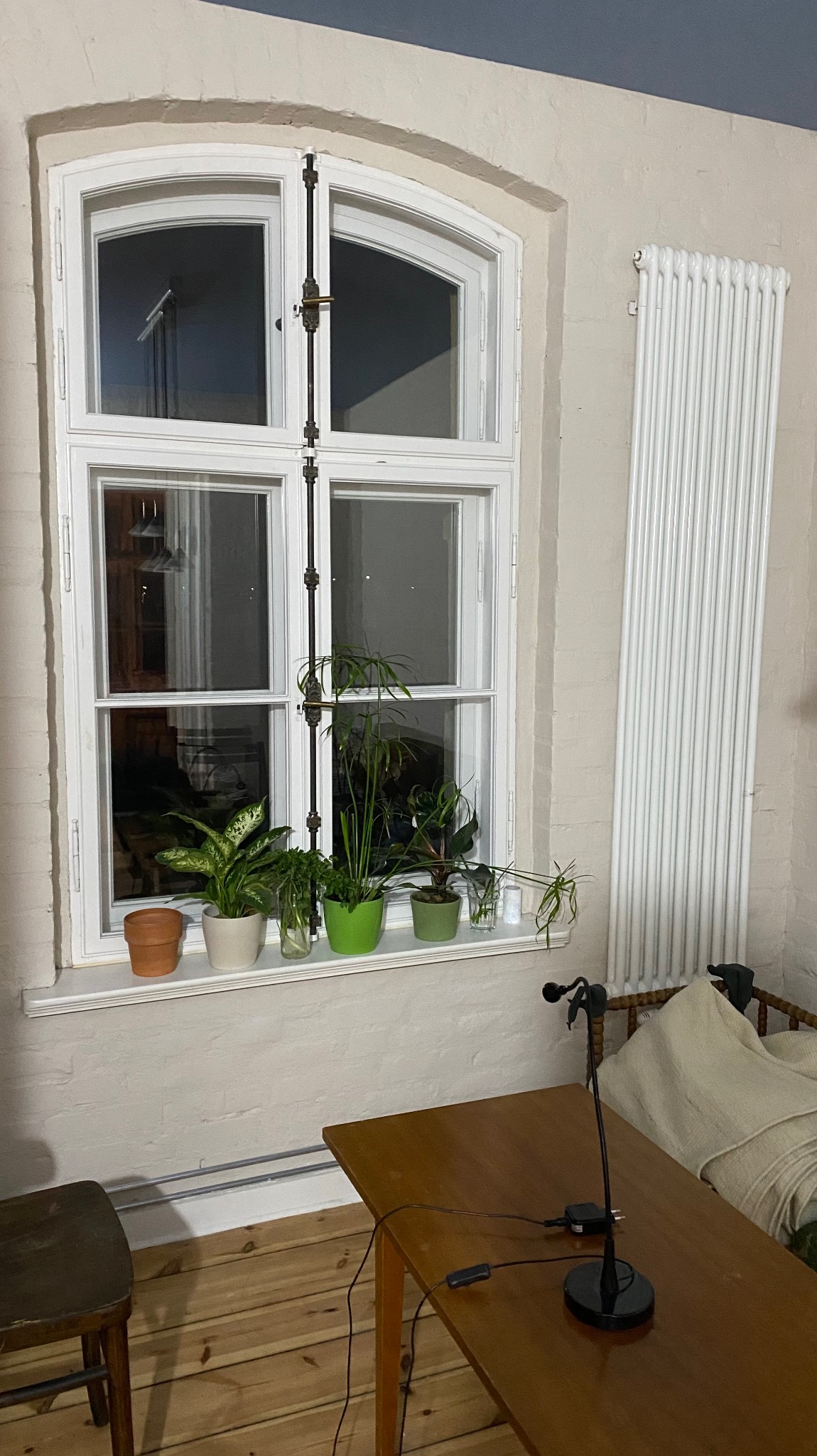 Białe, drewniane okno z roślinami doniczkowymi na parapecie. Widoczny grzejnik na ścianie i fragment drewnianego stołu z mikrofonem. Neutralne kolory ścian i podłogi.