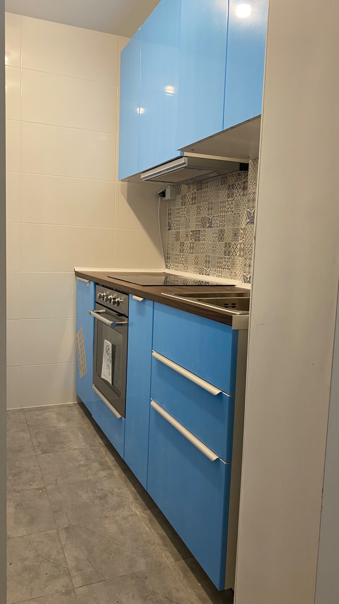 Błękitna zabudowa kuchenna z piekarnikiem i płytą grzewczą, wykończona brązowym blatem. Nad nią okap i dekoracyjne płytki. Szare płytki podłogowe.