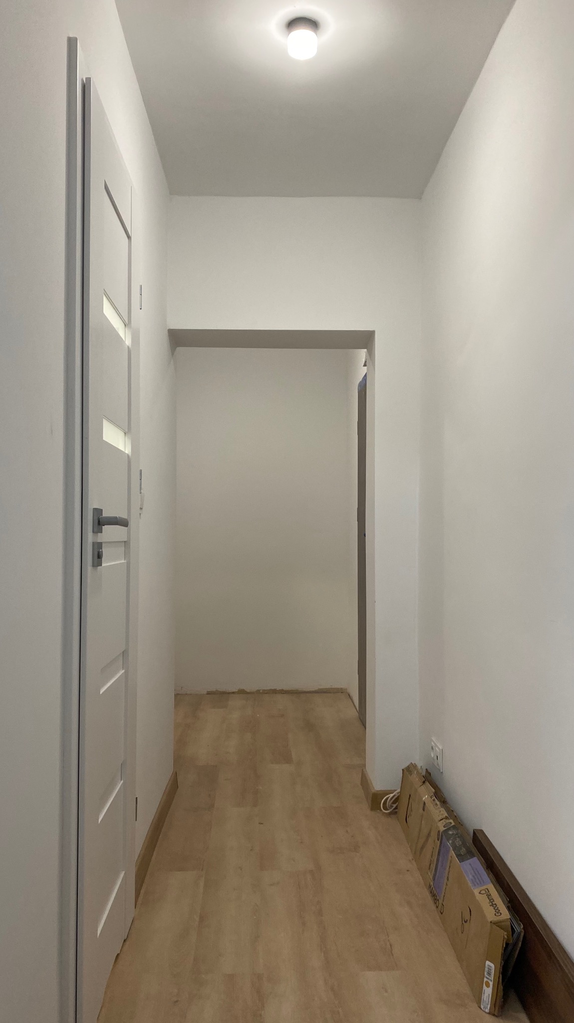 Jasny korytarz po remoncie z panelami podłogowymi i świeżo pomalowanymi ścianami. Widoczne kartony po panelach przy ścianie. Minimalistyczny design.