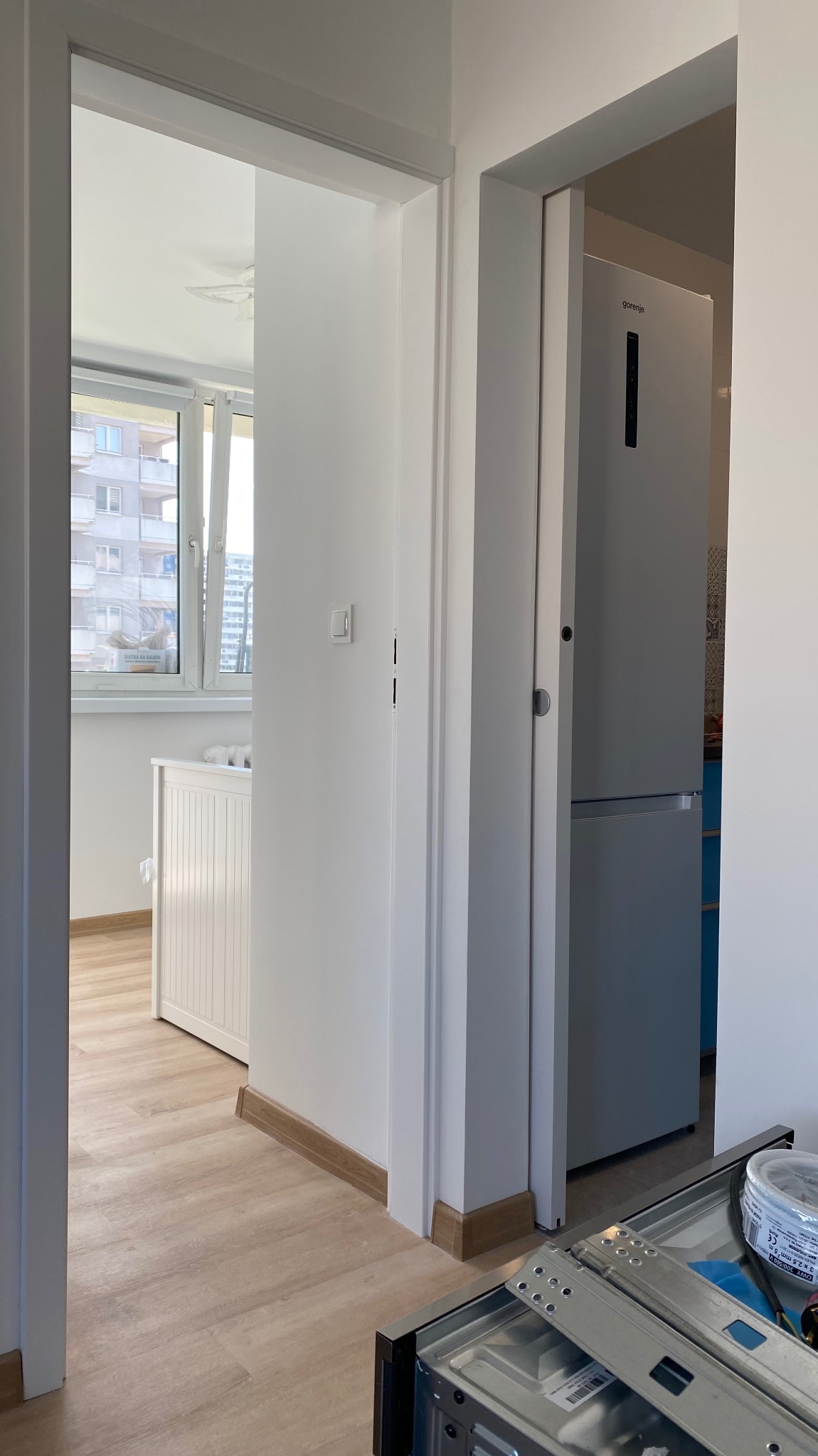 Białe, odnowione wnętrze mieszkania z widokiem na okno i zabudowaną lodówkę. Widoczne listwy przypodłogowe i fragment niedokończonej zabudowy meblowej.
