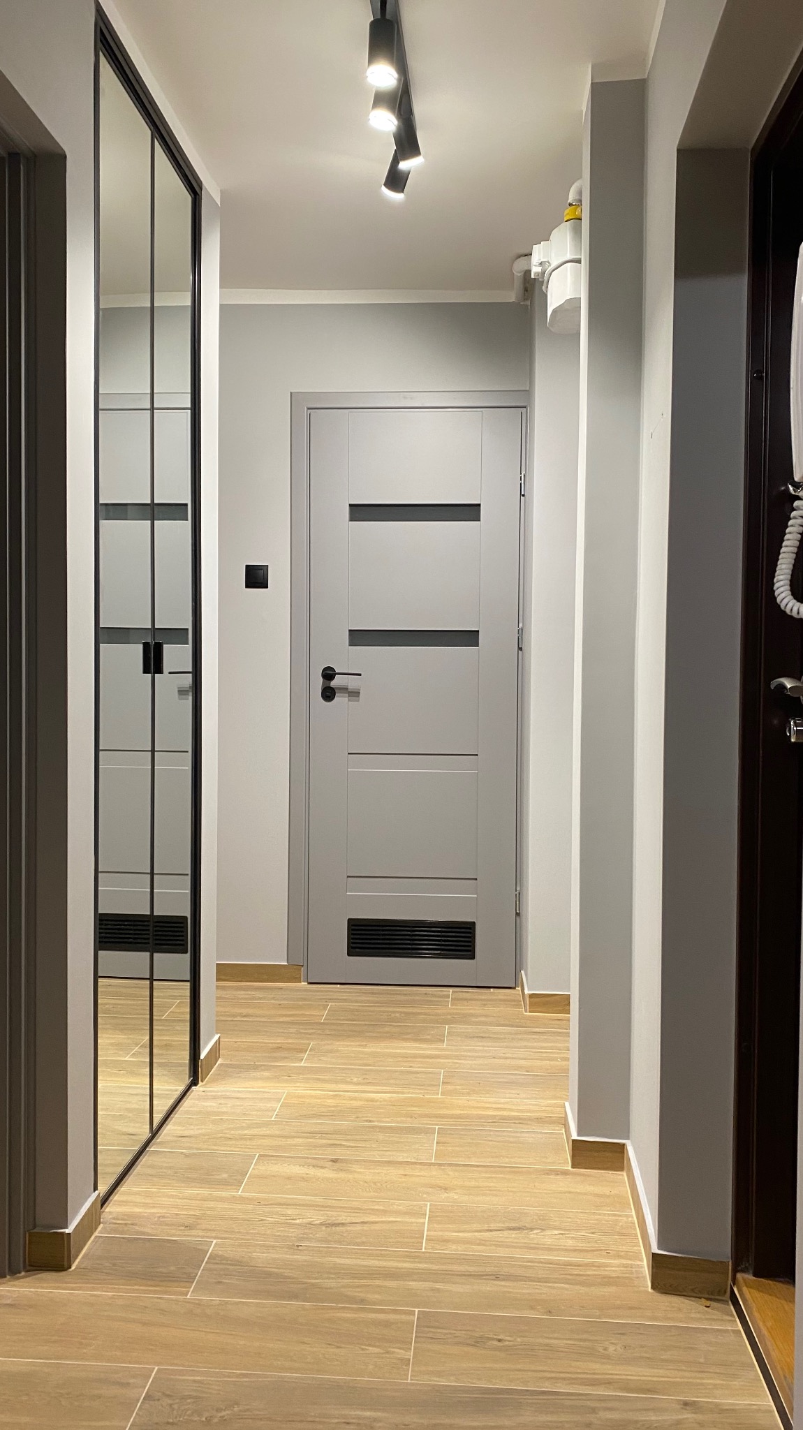 Wykończony korytarz w Katowicach: drewnopodobne płytki, szara ściana, drzwi wewnętrzne z przeszkleniami i lustrzana szafa w zabudowie. Nowoczesne oświetlenie.