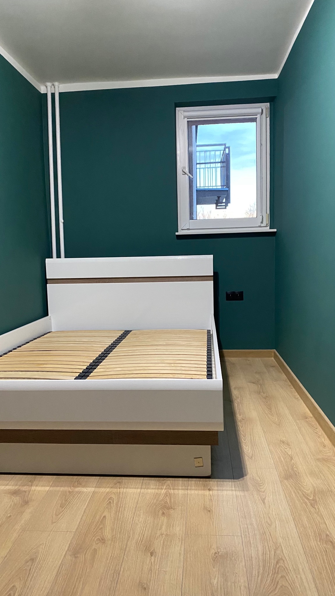 Nowoczesna sypialnia z drewnianą podłogą i ścianami w odcieniach zieleni. Białe łóżko z drewnianym stelażem, okno z widokiem na balkon. Minimalistyczny design.