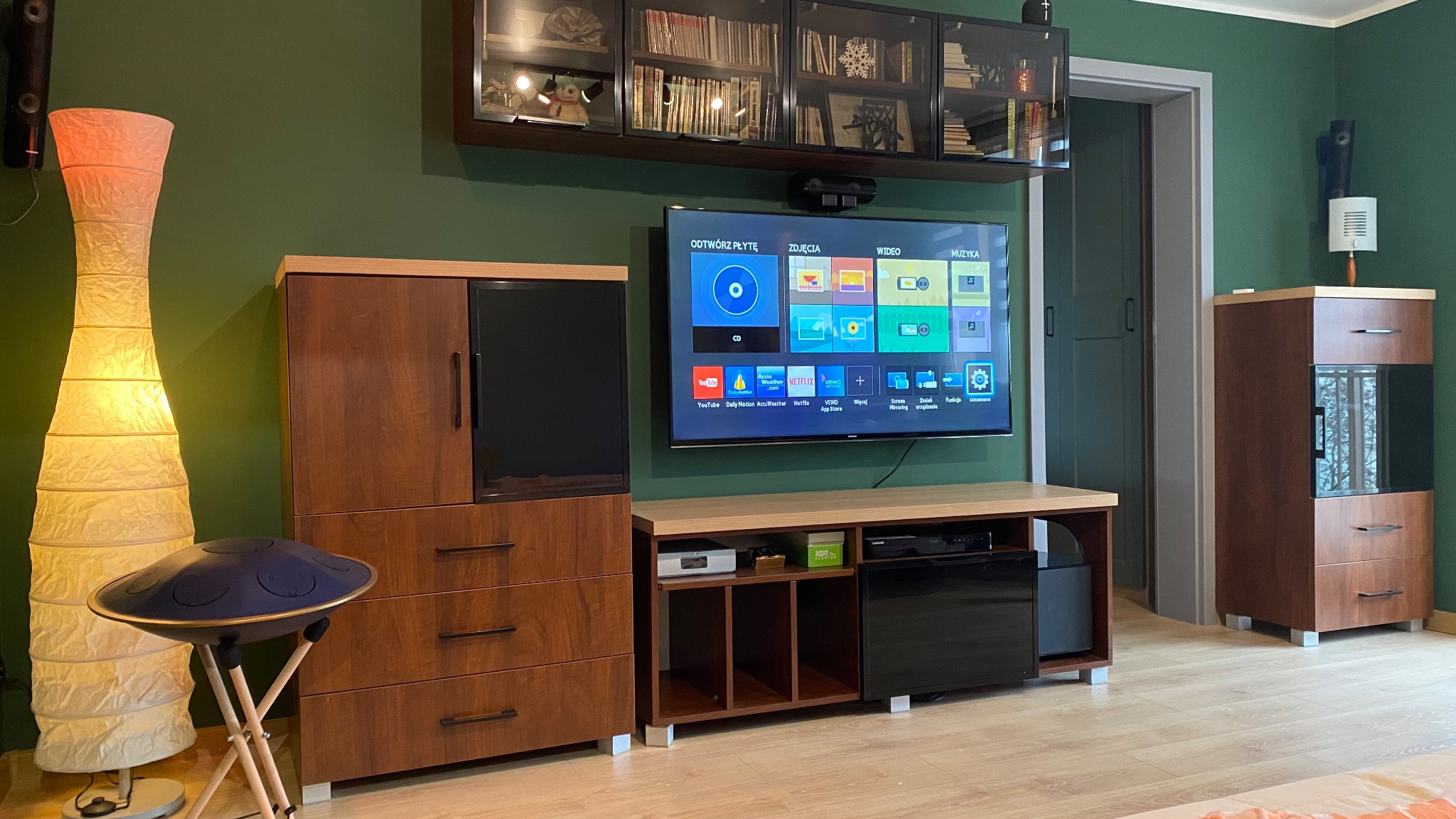 Nowoczesny salon z zielonymi ścianami, drewnianymi meblami, telewizorem i lampą. Widoczny hang drum na statywie. Minimalistyczny design, ciepłe oświetlenie.