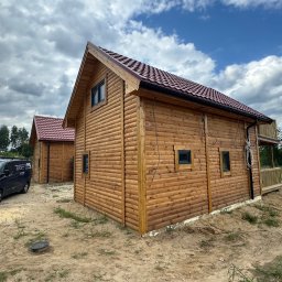 K&M NORD HAUS Szkieletowe Domy Skandynawskie - Dwa identyczne domki z bali z czerwonymi dachami, widoczne z zewnątrz, z niebieskim vanem z logo firmy w tle i częściowo odsłoniętą ziemią wokół.