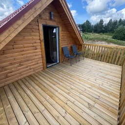 K&M NORD HAUS Szkieletowe Domy Skandynawskie - Drewniany domek z tarasem i dwoma krzesłami na tle zieleni i błękitnego nieba. Widok z góry, podkreślający strukturę drewna i przestrzeń tarasu.