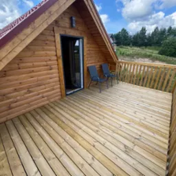 Drewniany domek z tarasem i dwoma krzesłami na tle zieleni i błękitnego nieba. Widok z góry, podkreślający strukturę drewna i przestrzeń tarasu.