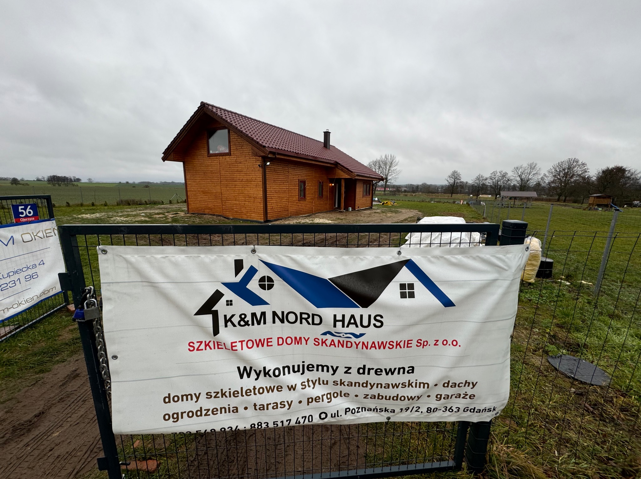 Gotowy, parterowy dom z brązowego drewna z ciemnoczerwoną dachówką, widziany zza ogrodzenia z banerem reklamowym firmy budującej domy w stylu skandynawskim, na tle pochmurnego nieba i zielonego pola.