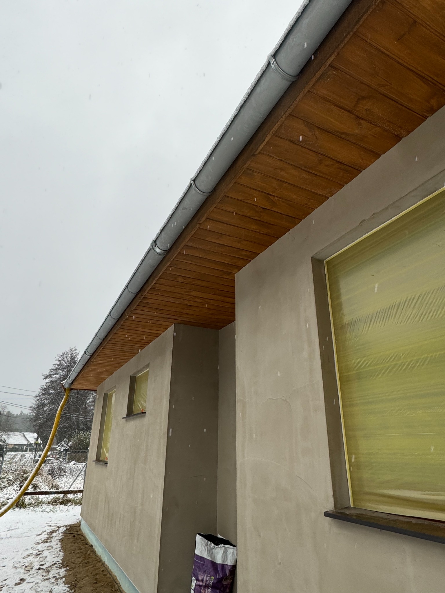 Surowy stan elewacji domu jednorodzinnego z drewnianą podbitką dachową i orynnowaniem, okna zabezpieczone folią, widoczny padający śnieg.