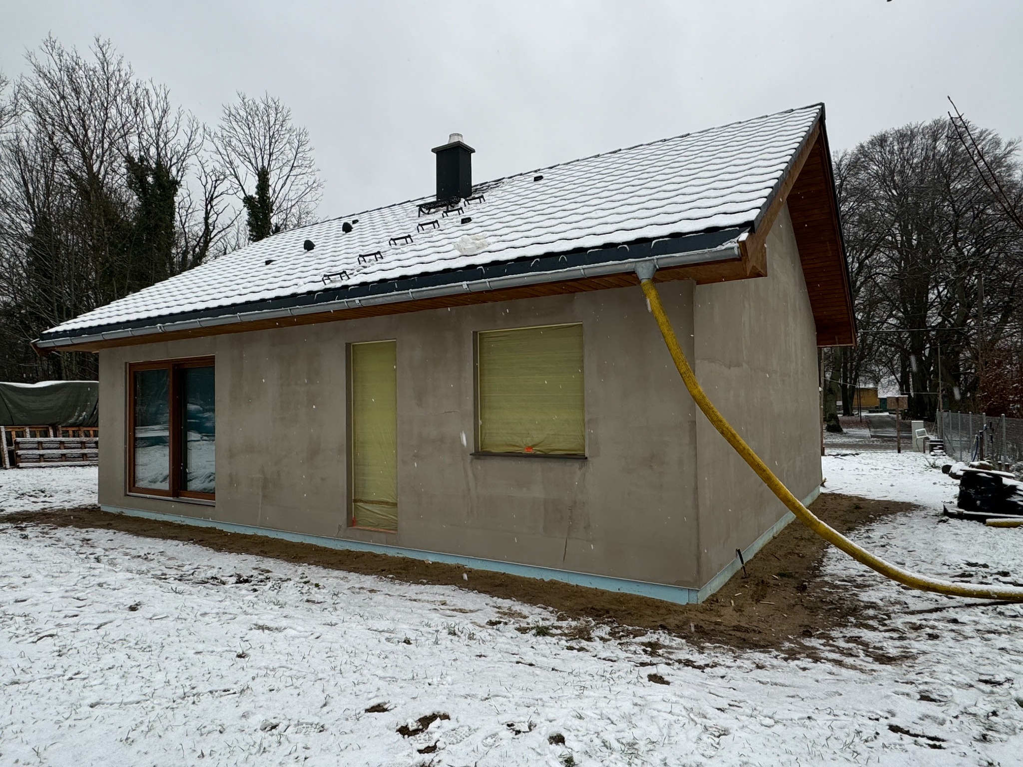 Niedokończony dom szkieletowy w zimowej scenerii, z oknami zabezpieczonymi folią i widocznym ociepleniem fundamentów, żółty wąż przy elewacji.