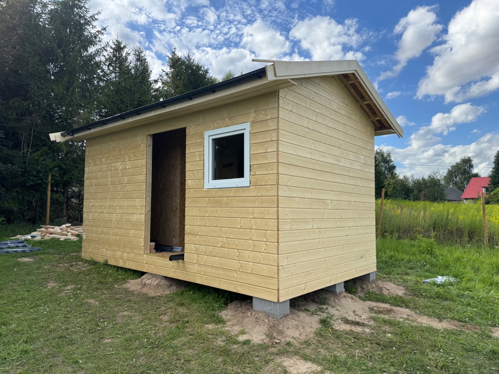 Wznoszenie małego, drewnianego domku letniskowego na betonowych bloczkach, z widocznym oknem i otwartym wejściem, na tle zieleni i błękitnego nieba.