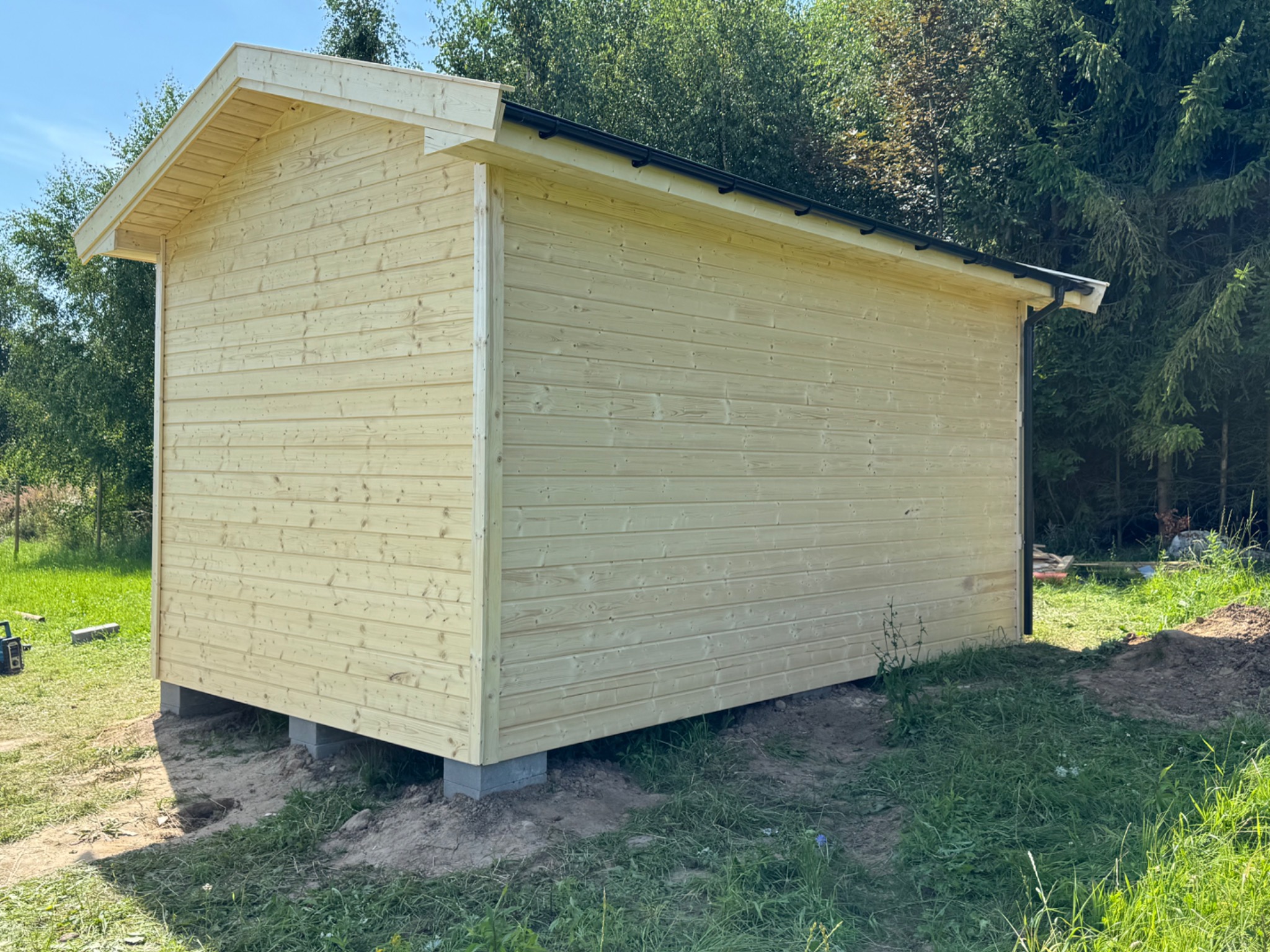 Gotowy mały drewniany budynek z czarną rynną, osadzony na betonowych blokach, otoczony zielenią. Widok z boku, w tle drzewa.